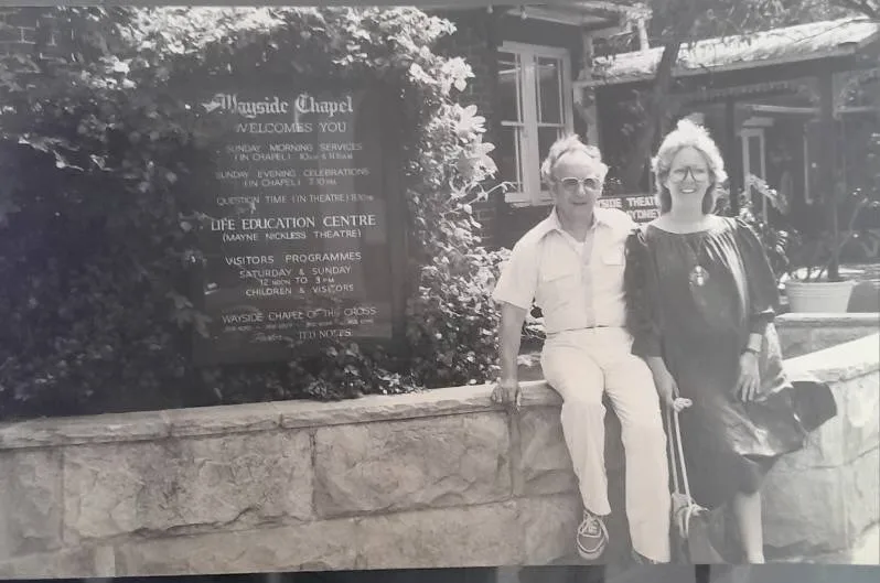 An old black and white image of Mandy as a young woman sitting on the wall outside the Wayside chapel with Rev Ted Noffs.