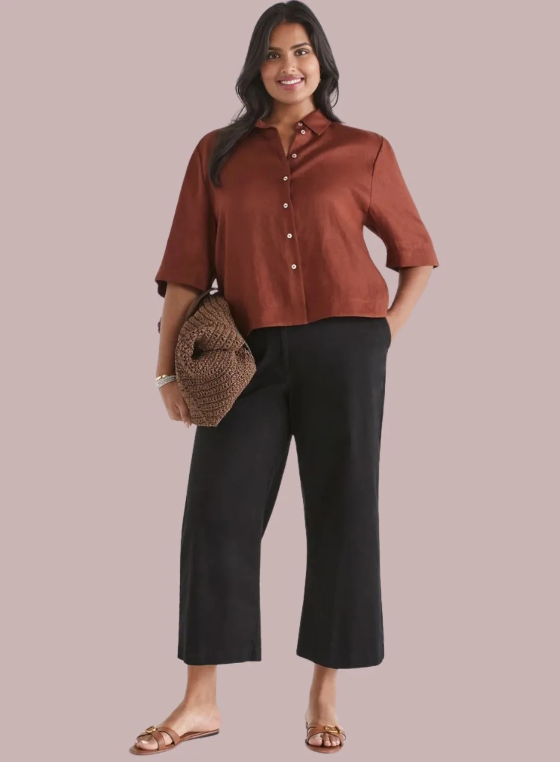 A woman modeling a rust-colored short-sleeve button-down shirt and black cropped wide-leg trousers, holding a woven brown clutch.