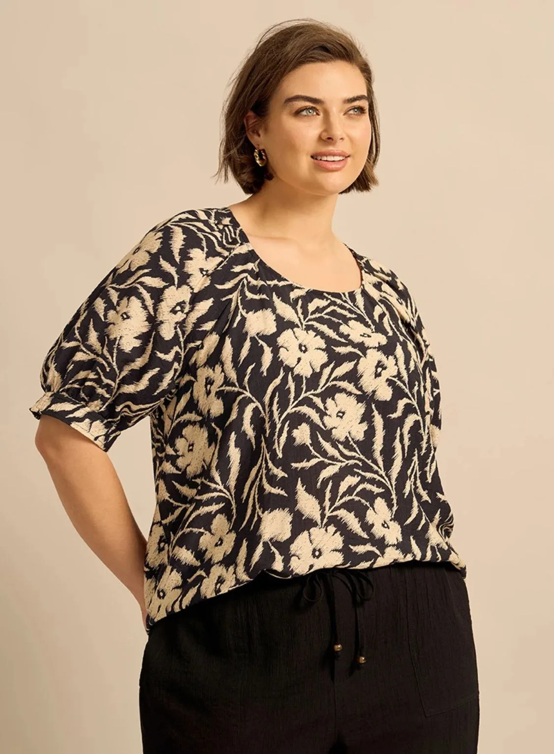 A woman with a bob hairstyle wearing a black blouse with a cream floral pattern and black drawstring trousers from Suzanne Grae.