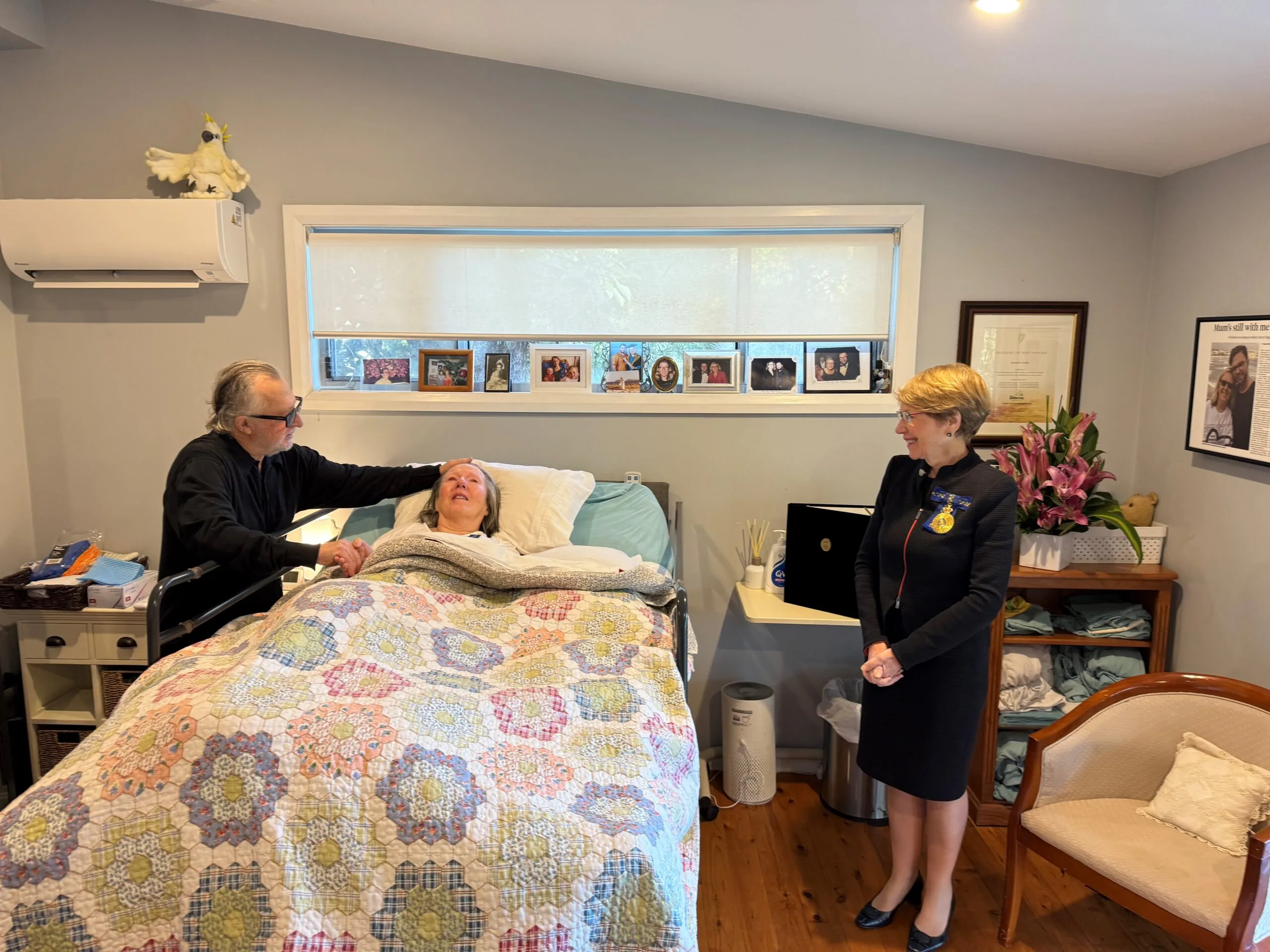Wesley leans over Mandy who lies in bed covered by a patchwork quilt. On the other side, stands the Governor in a navy suit.
