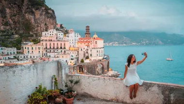A woman in a white dress sits on a stone ledge taking a selfie on her smartphone, with the coastal town of Atrani on Italy's Amalfi Coast and the blue ocean in the background.