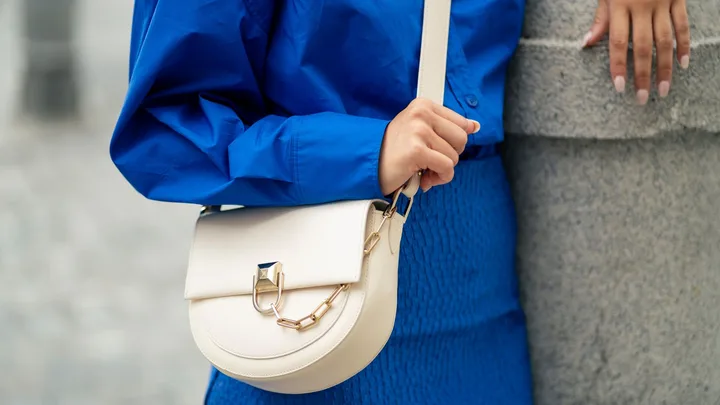 Close-up of a person wearing a vibrant blue button-down shirt and skirt, showcasing a cream-coloured crossbody saddle bag with gold hardware.