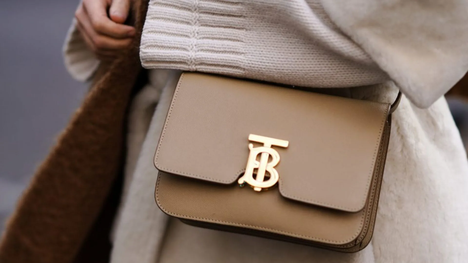 A close-up street style photograph focusing on a smooth tan leather Burberry TB Shoulder Bag. The structured bag features a prominent interlocking gold-tone "TB" monogram clasp and is styled against a cosy, cream-coloured ribbed knit sweater and a plush cream coat. Featured in AWW's heritage luxury brands article.
