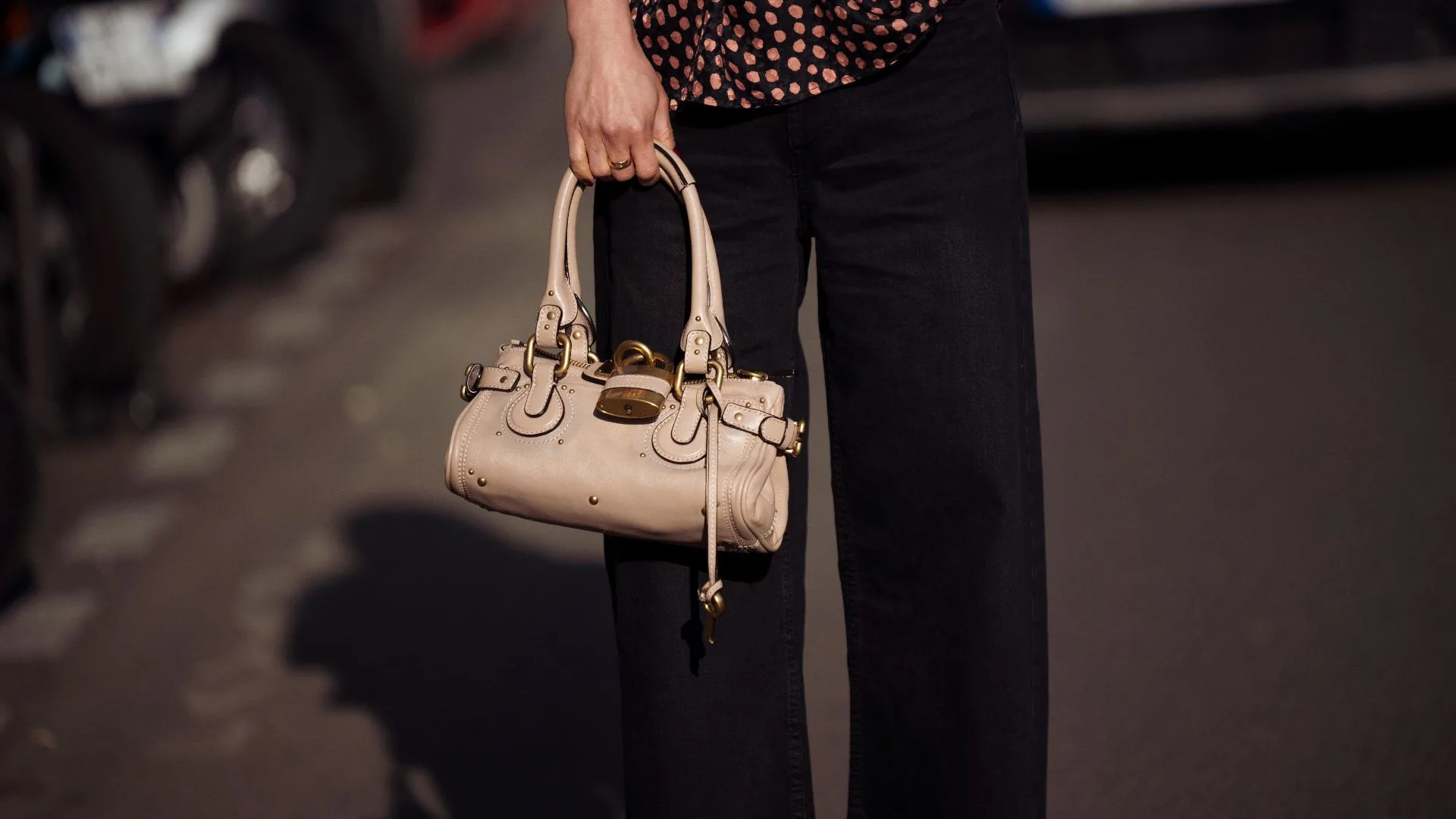 A close-up street style photograph from Paris Fashion Week showing a person from the waist down wearing black wide-leg trousers and a ruffled polka-dot blouse. They are holding a beige leather Chlo&eacute; Paddington handbag, showcasing its iconic slouchy shape and prominent brass padlock hardware. Featured in AWW's heritage luxury brands article.