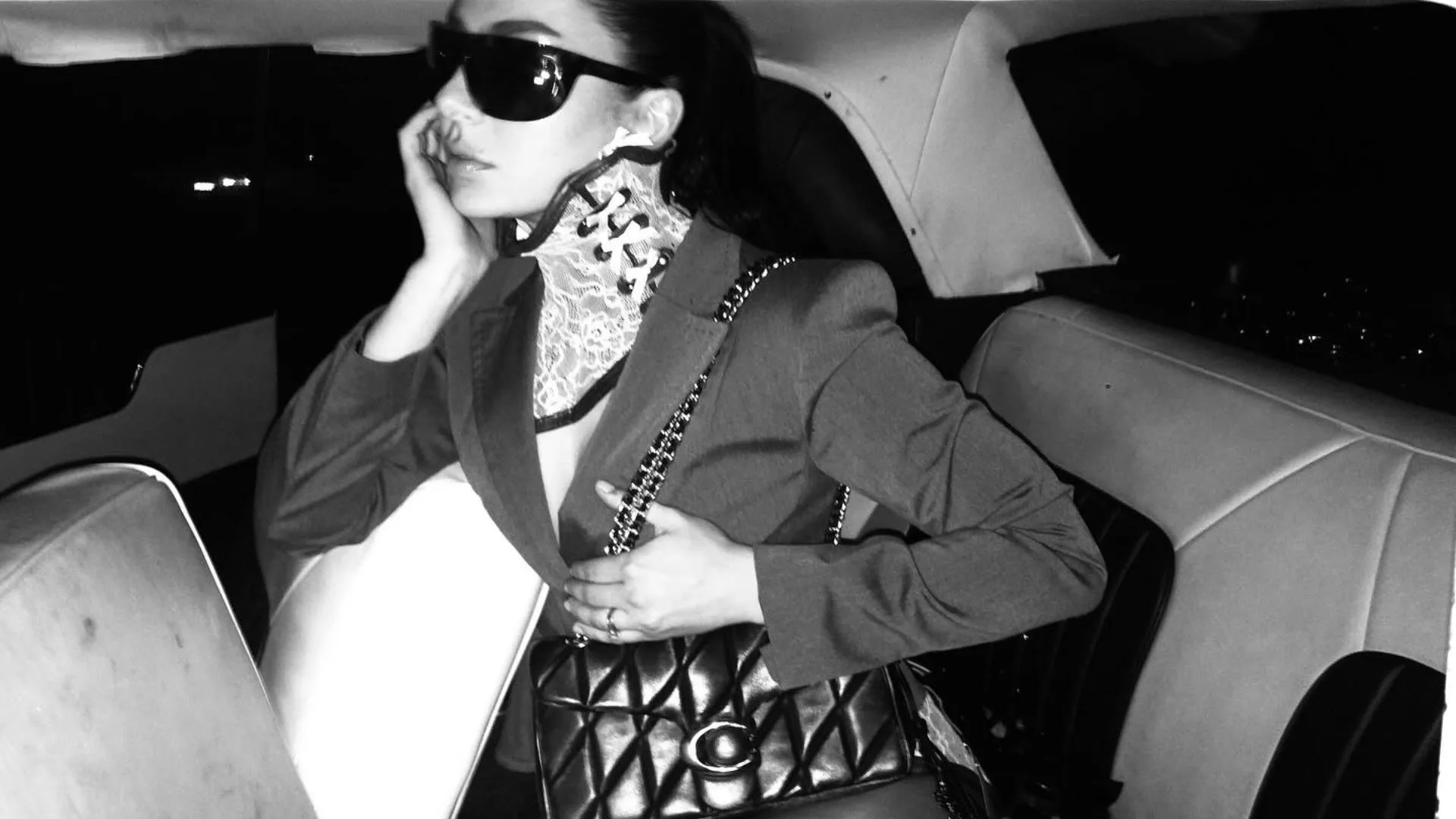 Black and white photograph from the "Rock Music" music video featuring Charli XCX in the backseat of a car. She wears dark sunglasses, a structured blazer, and a white lace choker, while holding a black Coach Quilted Tabby 26 handbag with a chain strap over her shoulder. Featured in AWW's heritage luxury brands article.