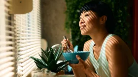 A smiling woman eating a healthy breakfast bowl with blueberries near a sunlit window with blinds.