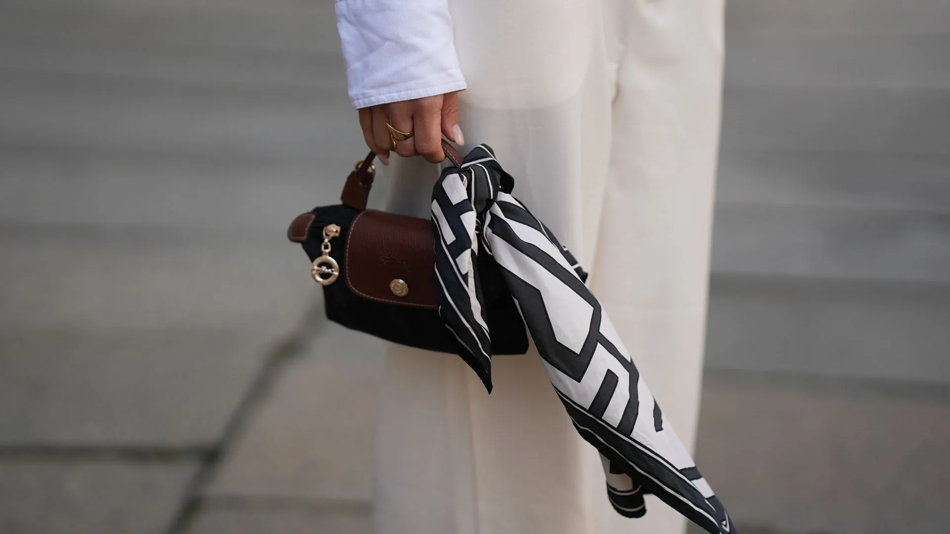 Close-up street style photograph of a person holding a small black nylon Longchamp "Le Pliage Mini Bag" with brown leather top handles and a gold charm. A black and white graphic printed scarf is tied around the handle, contrasted against cream wide-leg trousers.