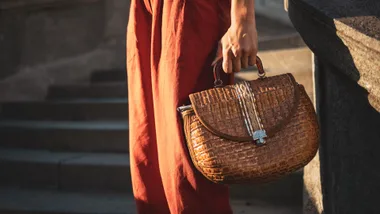 A close-up of a woman in rust-coloured trousers holding a brown woven basket bag, showcasing style inspiration for luxury handbags under $1000.