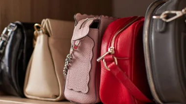 Close-up editorial shot of multiple designer handbags on a wooden shelf, showcasing different colors and textures, with the sharpest focus on a pastel pink pebbled leather bag featuring scalloped trim.