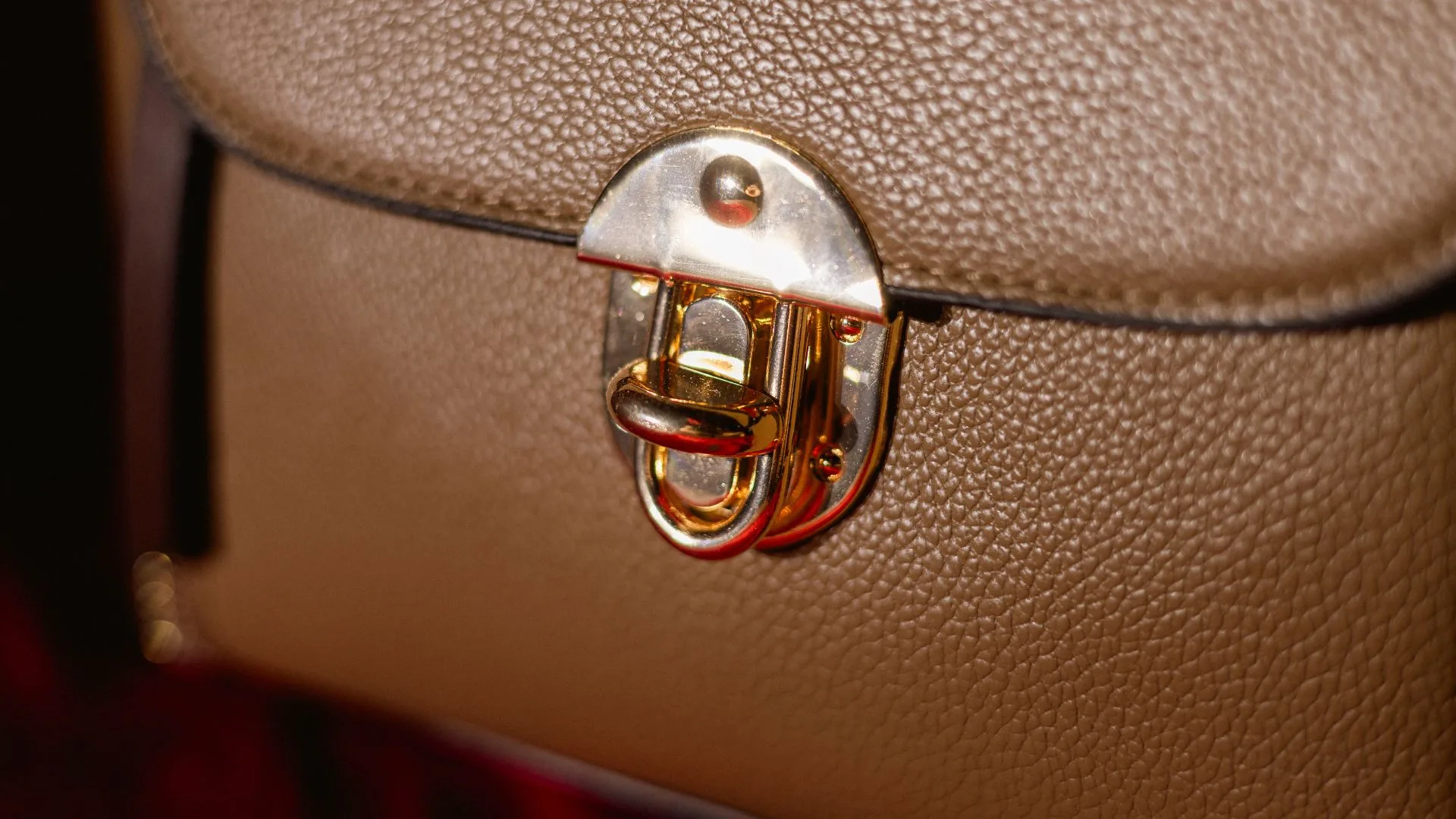 A detailed macro shot of a polished gold clasp on a textured tan leather handbag. The warm lighting emphasises the craftsmanship of the pebbled leather bag, highlighting the high-end material and refined design.