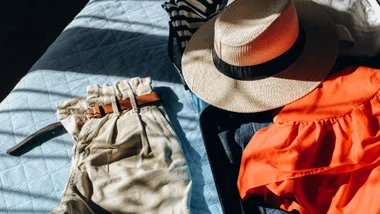 Summer holiday clothes, a straw fedora hat and cream shorts laid out on a light blue quilted bed next to an open suitcase demonstrating smart travel packing hacks.