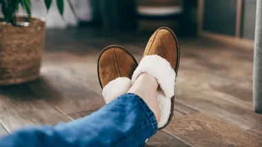 A close-up lifestyle shot of a person relaxing on a wooden floor, wearing a pair of classic tan suede scuff slippers with fluffy white sheepskin lining. The person is wearing blue jeans, with their feet kicked up comfortably in a cosy indoor setting.