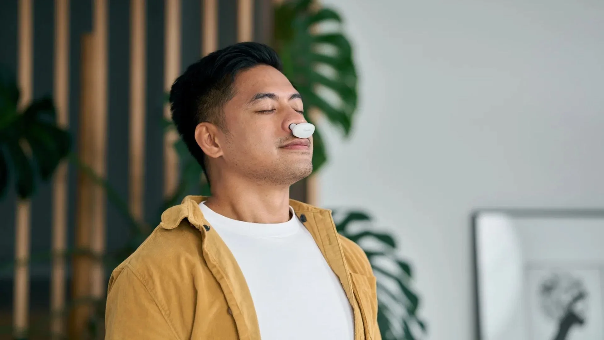 A man with his eyes closed wearing a small, white Goodair Nosebuds device in his nose, looking relaxed in a modern room with indoor plants.