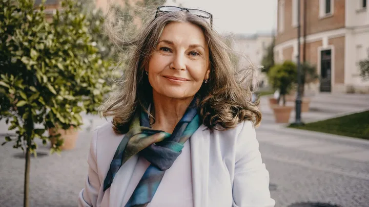 Close-up portrait of a middle-aged woman with thick, glossy grey hair smiling on a sunny street, wearing a light blazer and silk scarf.