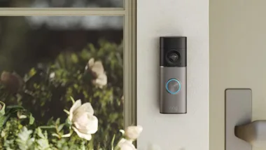 A Ring Video Doorbell mounted on a light-coloured wall next to a front door window. Featured in smart doorbells article on AWW.