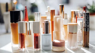 A collection of makeup and skincare products, including foundation, lip gloss and serums, arranged on a vanity in natural sunlight.