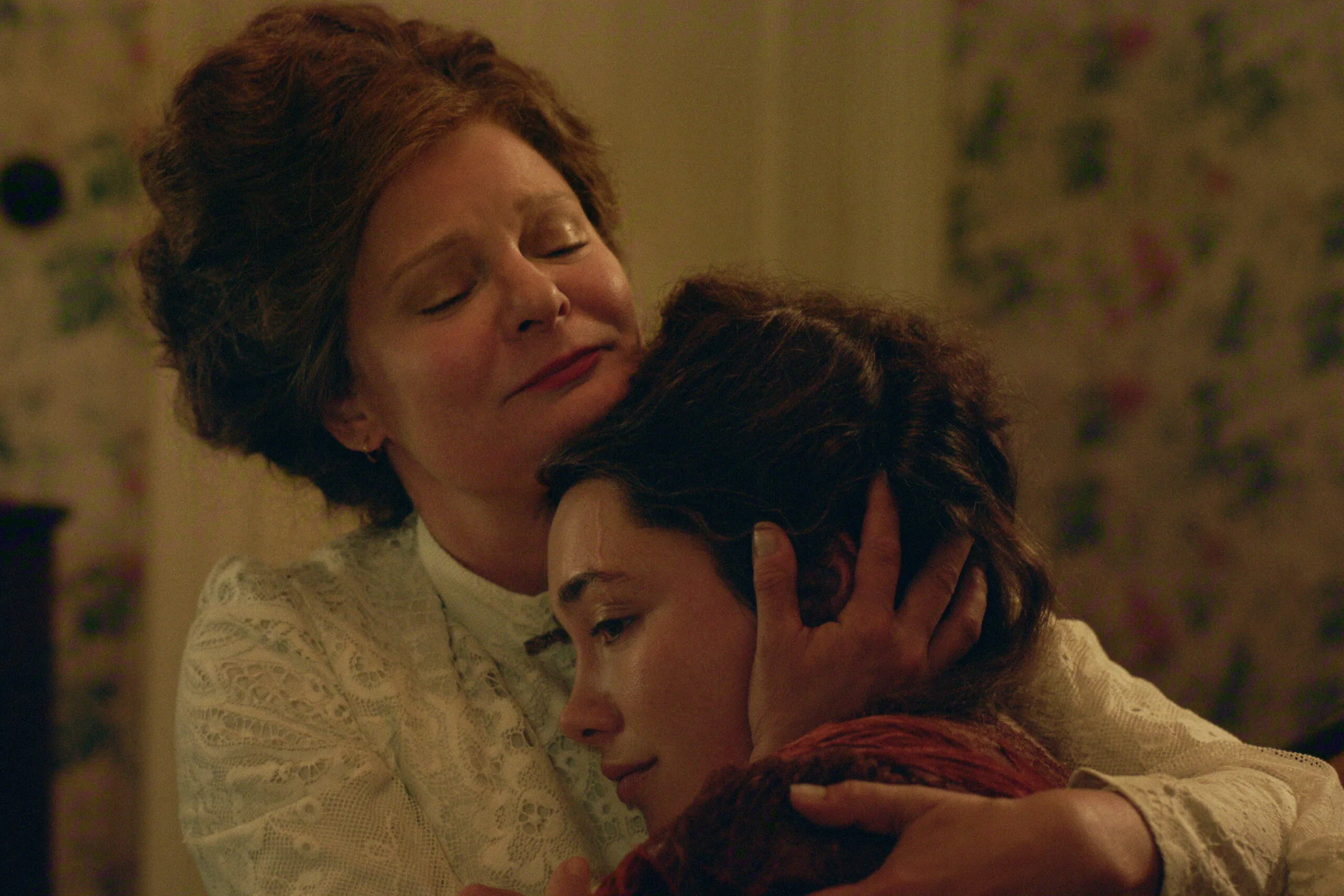"Two women in vintage clothing share an emotional embrace in a warmly lit, floral-wallpapered room, suggesting comfort and support in a historical or period setting."