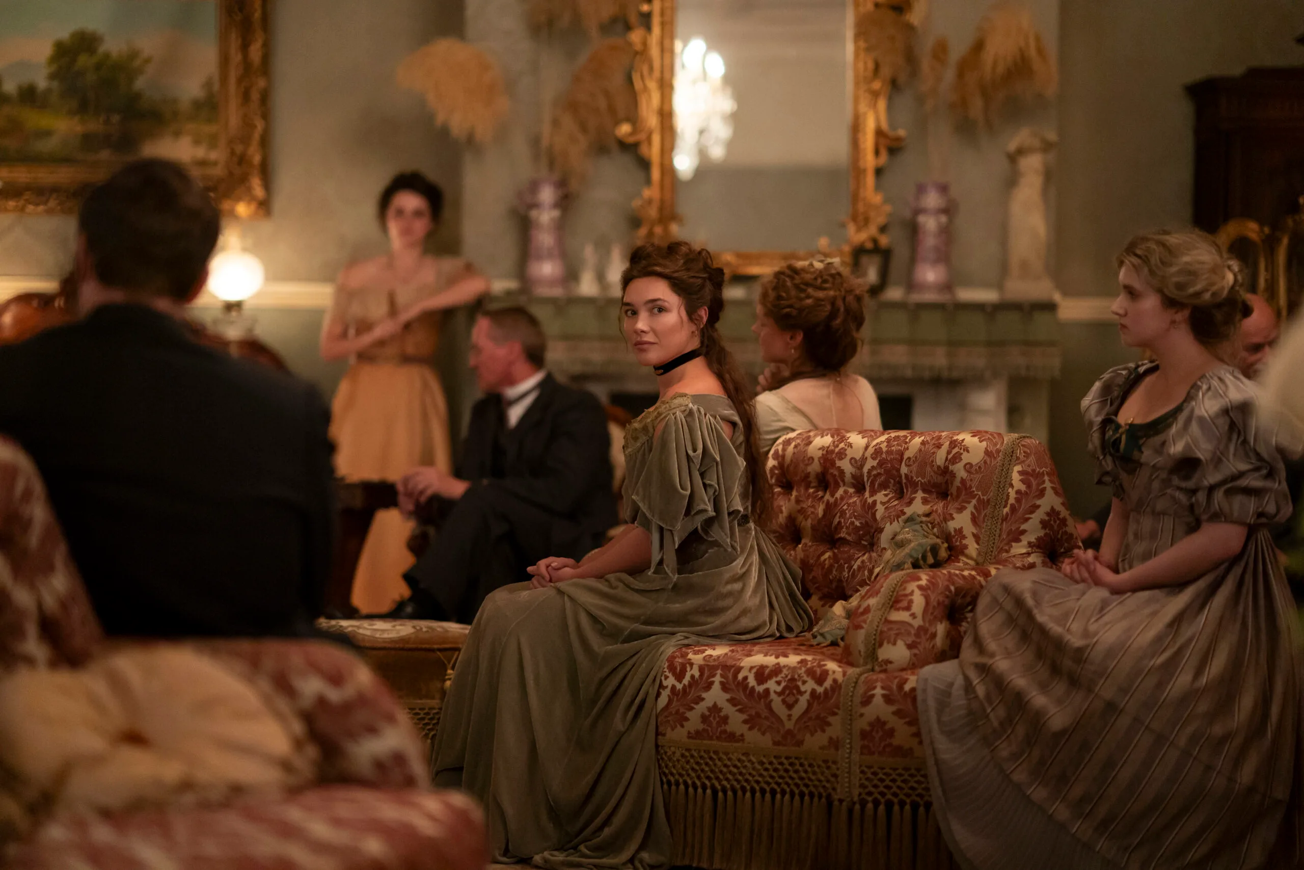 "A group of people in period clothing sit in an ornate, vintage living room with elegant furniture, antique decorations, and a large mirror above the fireplace, engaged in conversation or listening."