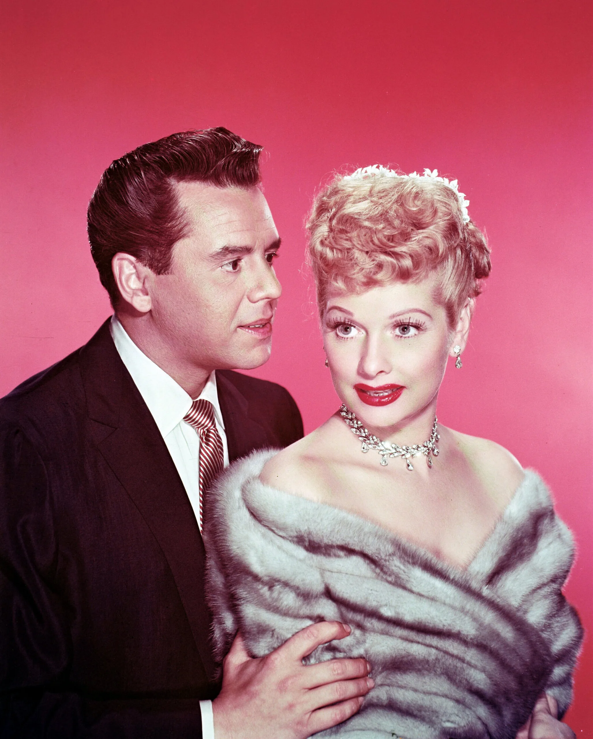Desi Arnaz (1917-1986), Cuban musician and actor, with Lucille Ball (1911-1989), US comedian and actress, wearing a grey fur stole, in a studio portrait, against a red background, circa 1955. (Photo by Silver Screen Collection/Getty Images)
