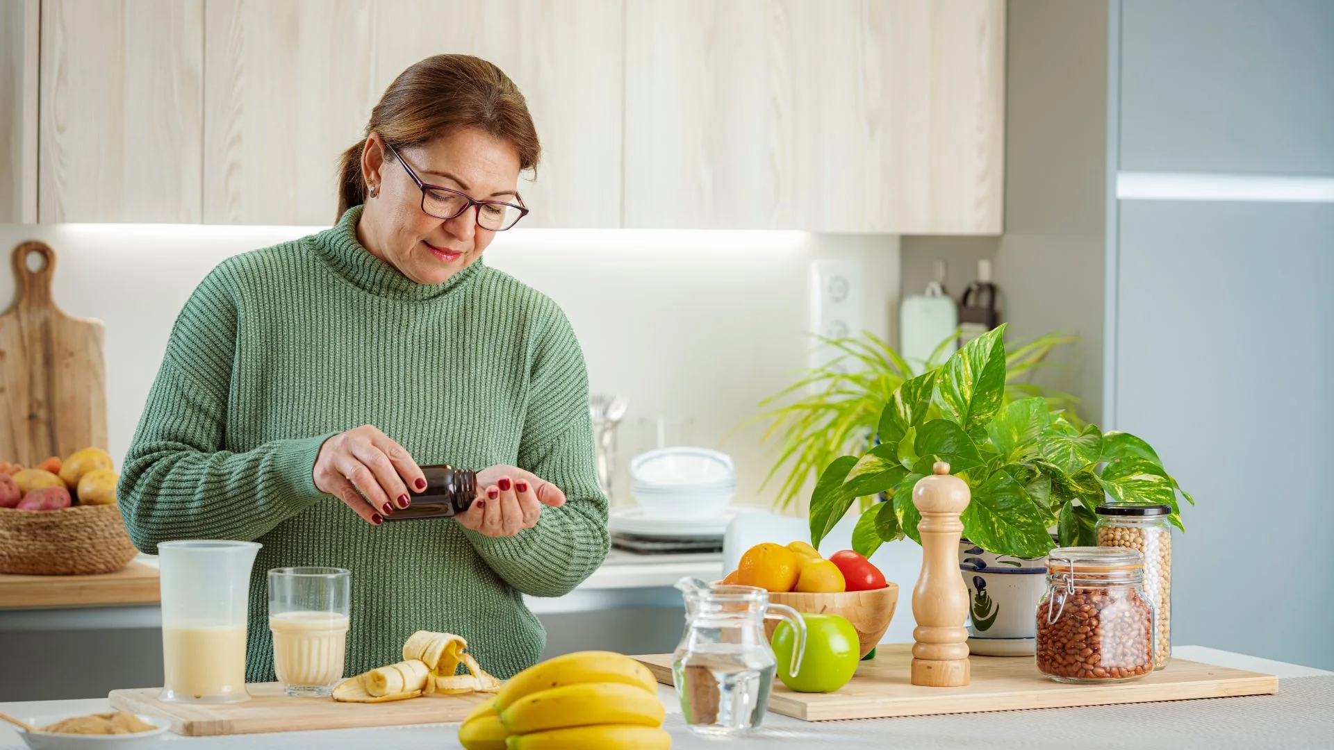A woman in a green sweater standing in her kitchen pouring supplement pills into her hand beside fresh fruit and a smoothie.