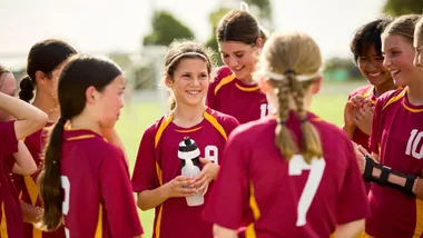 Young female sports team in matching maroon and yellow uniforms smiling and talking during a water break on a sunny field.