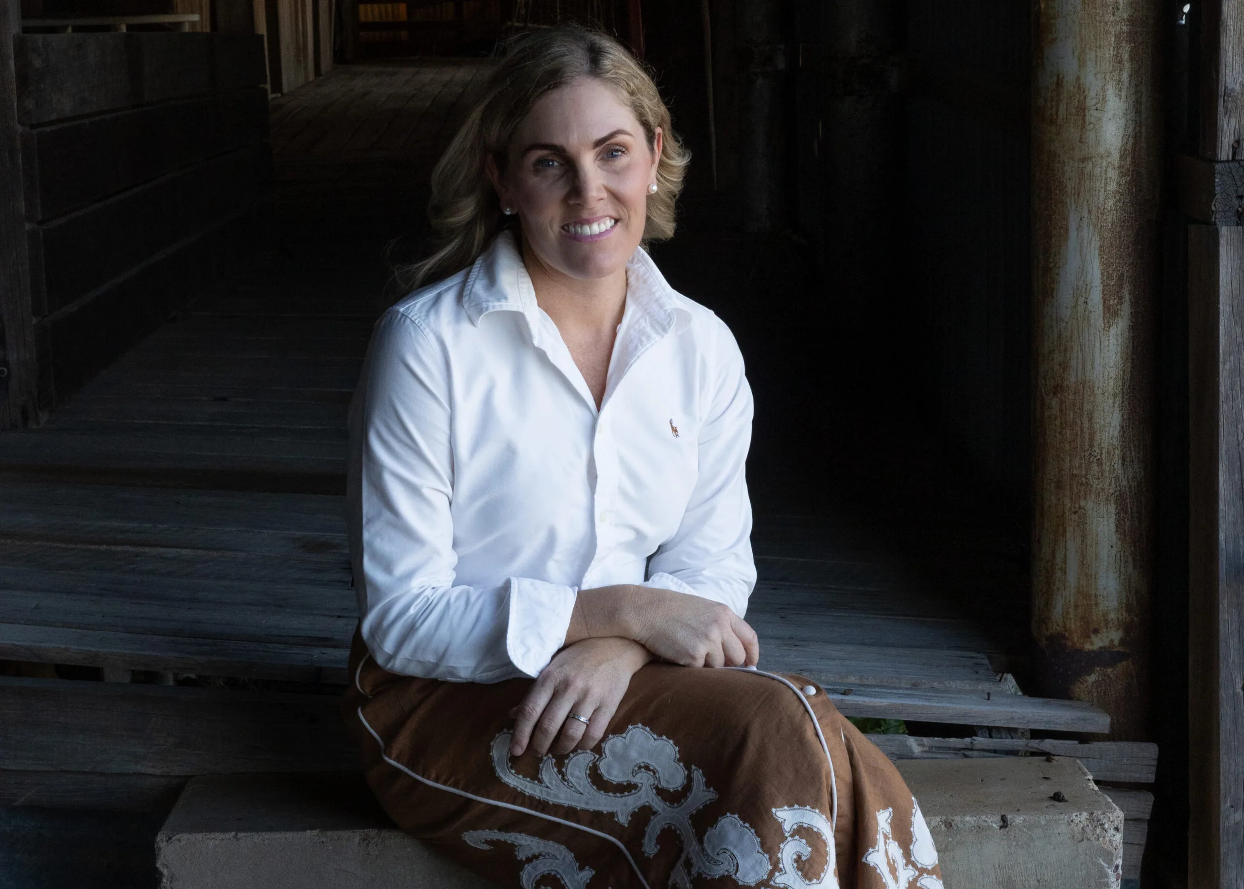 Angie sits in an old, wooden barn, She wears a white shirt and a brown skirt.