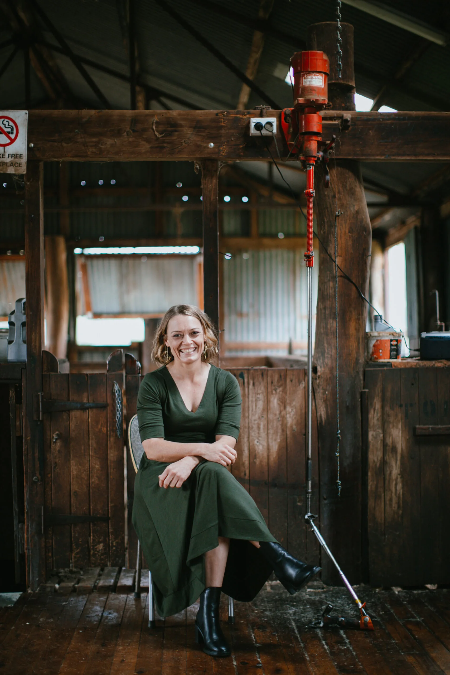 Carol wears a dark green dress and sits on a chair in an old wooden shearing shed.
