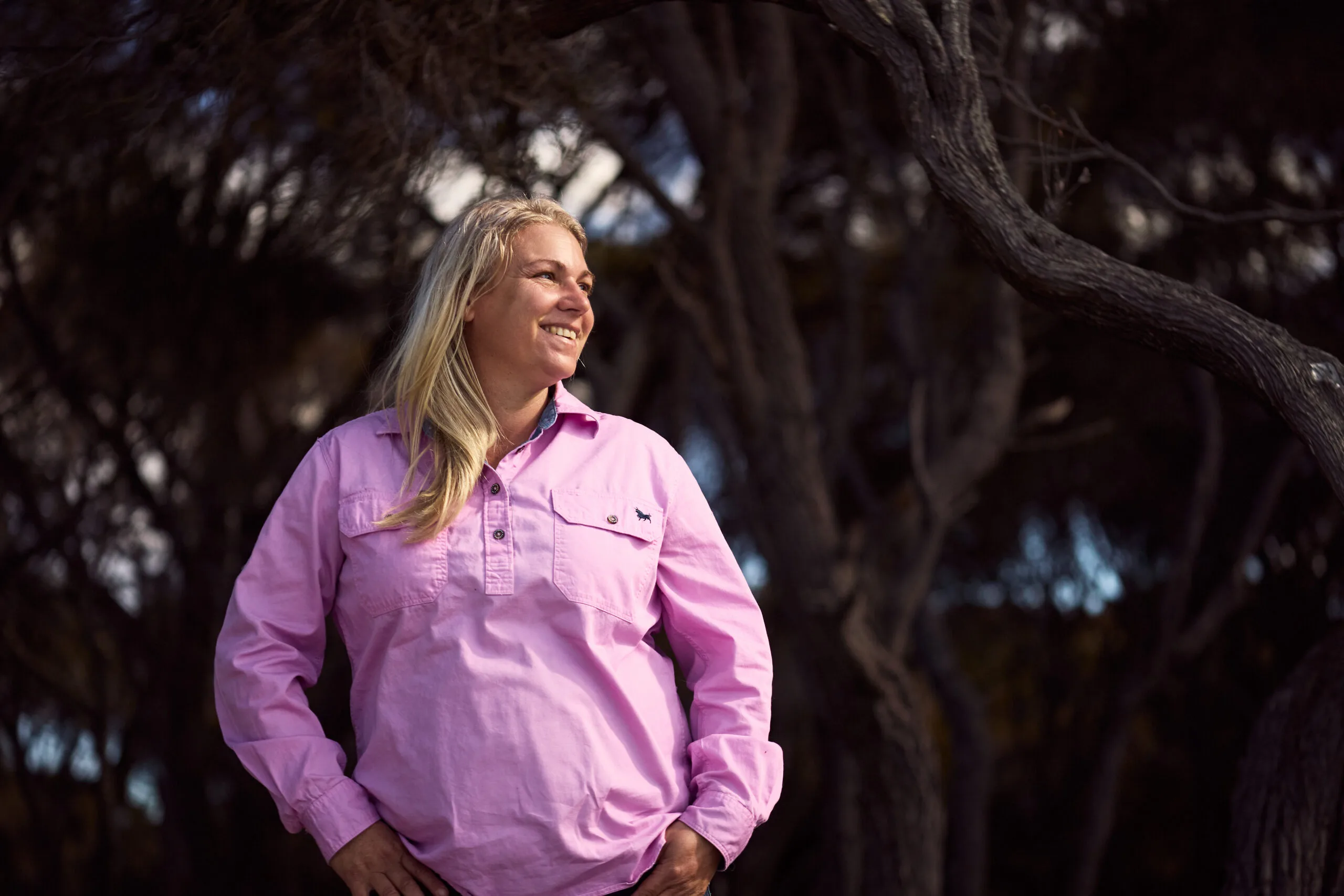 Amy stands in a paddock with trees behind her. She wears a pink farming shirt.