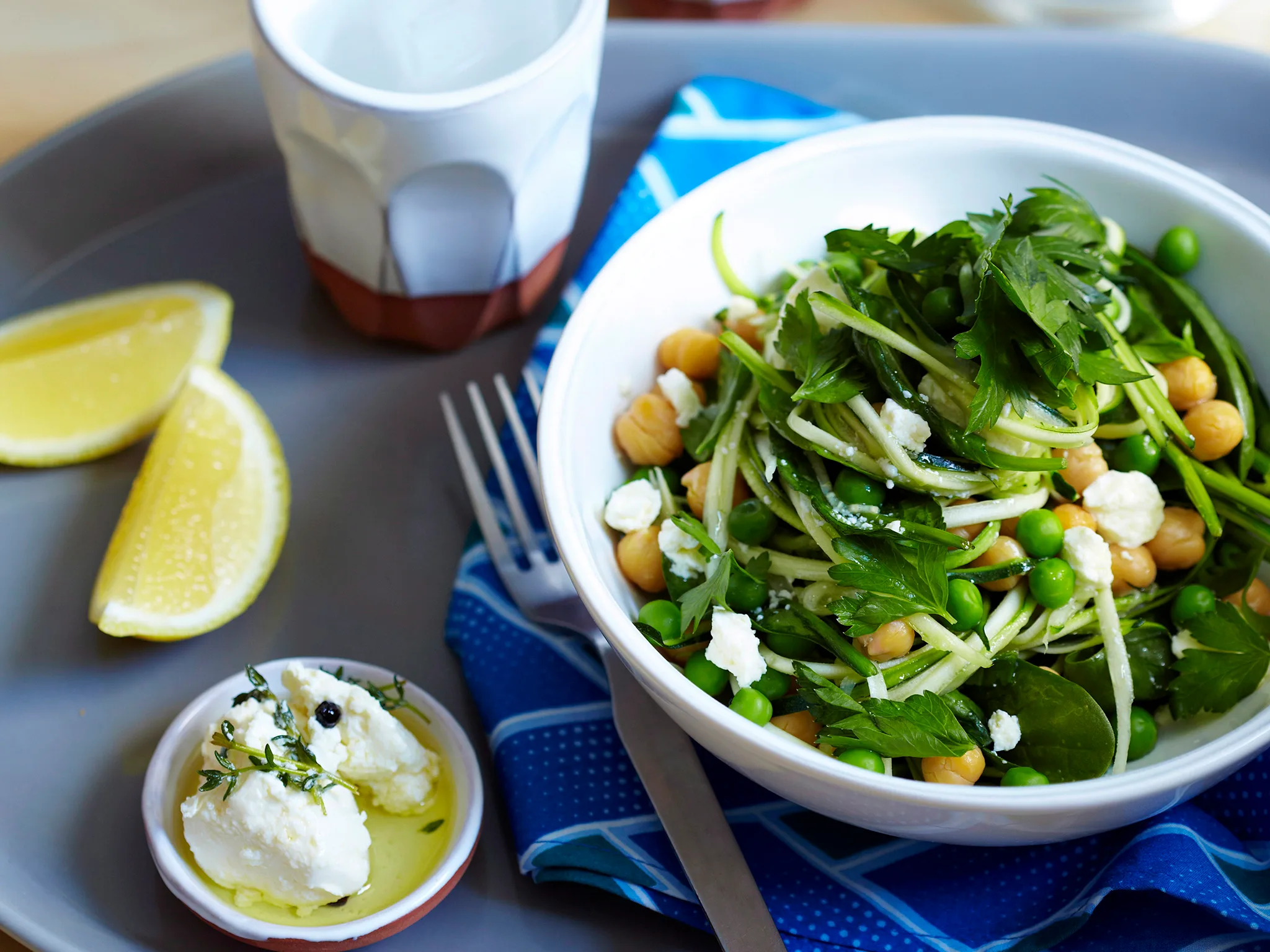 Bowl of zucchini noodles with chickpeas, peas, and feta on blue napkin, with lemon wedges and cheese nearby.