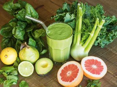 Green smoothie surrounded by spinach, kale, celery, grapefruit, lemon, avocado, mint, and pear on a wooden table.