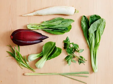 A selection of Asian greens on a wooden surface, including bok choy, lemongrass, daikon radish, and other leafy greens.