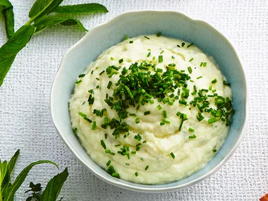 Bowl of creamy mashed potatoes garnished with chopped chives on a textured white background.