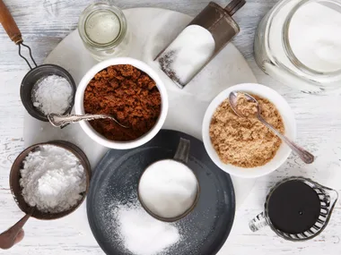 Assorted natural sweeteners in bowls and jars, including brown sugar, molasses, and powdered sugar, on a wooden table.