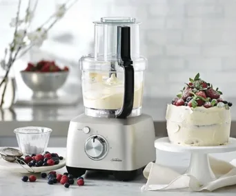 A Sunbeam food processor with a clear bowl and white contents, on a marble countertop with a cake and berries.