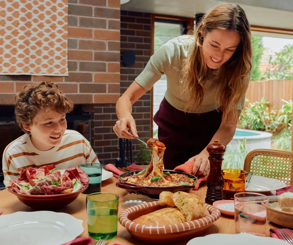 A smiling woman serves a cheesy, saucy dish from a cast-iron pan to a boy sitting at a table.