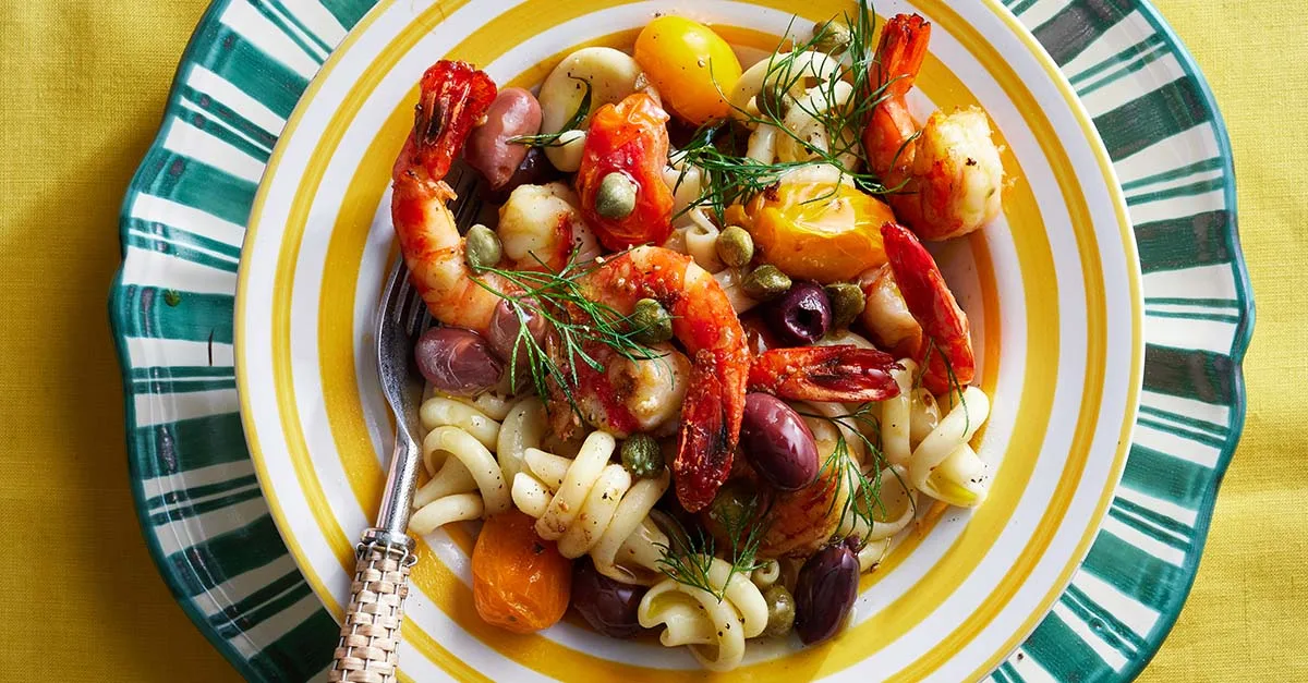 Colorful pasta salad with shrimp, cherry tomatoes, olives, and capers on a striped yellow and green plate.