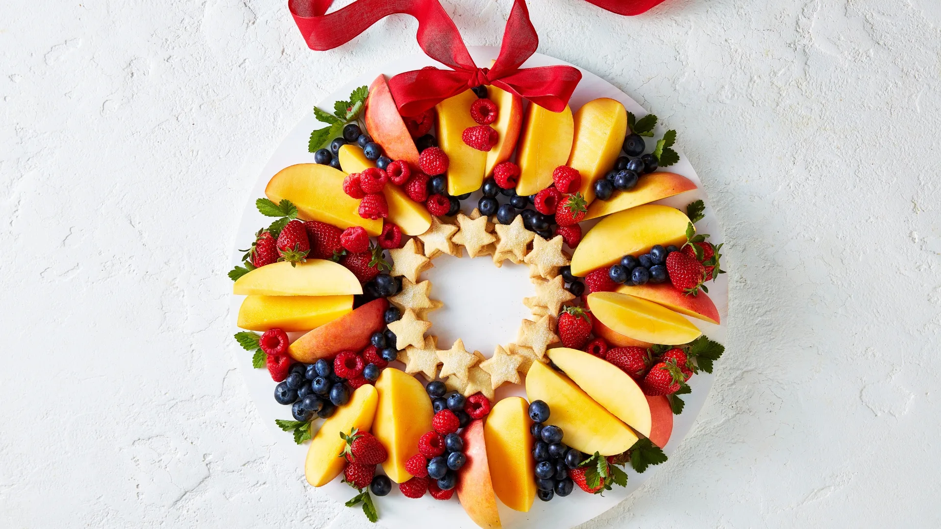Fruit wreath with apple slices, berries, and star-shaped cookies on a plate, topped with a red ribbon.