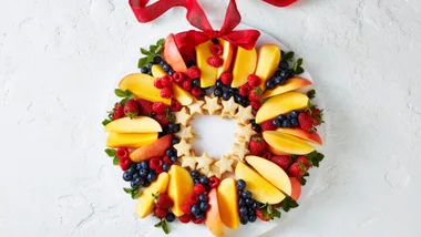 Fruit wreath with apple slices, berries, and star-shaped cookies on a plate, topped with a red ribbon.