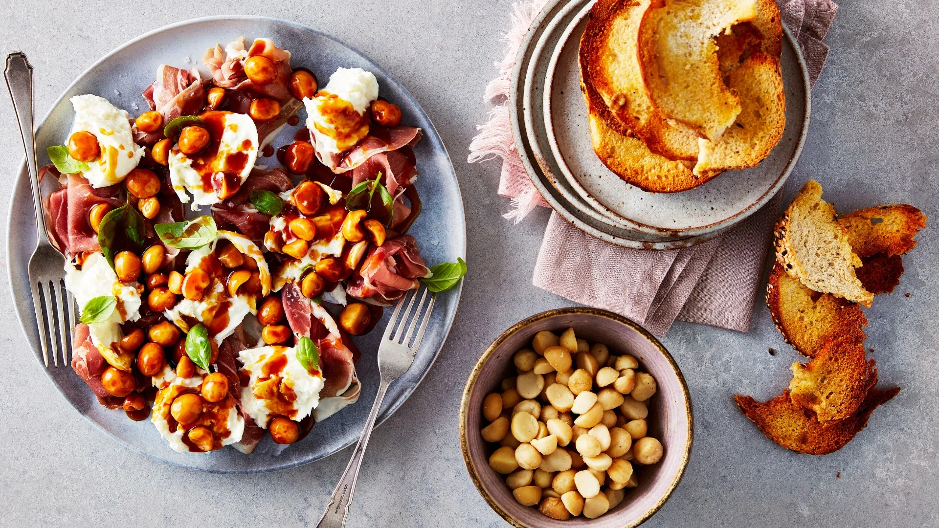 Prosciutto and cheese salad with roasted chickpeas, fresh basil, macadamia nuts, and crispy bread slices on a gray table.