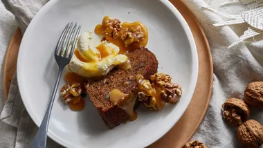 Slice of sticky date pudding with walnuts, ice cream, and caramel sauce on a white plate with a fork.