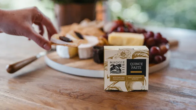 A hand holding cheese near a box of Maggie Beer quince paste on a wooden table with a cheese platter.