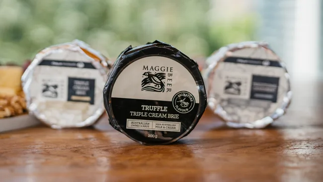 Maggie Beer Truffle Triple Cream Brie cheese package on a wooden table, with two blurred packages in the background.