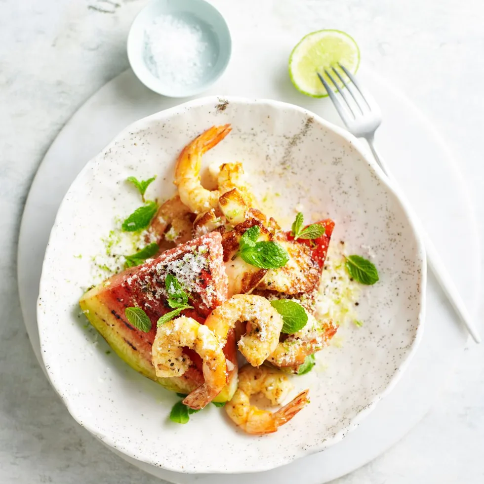 Watermelon, halloumi and prawn salad in a shallow white bowl with a lime half and a salt bowl beside it