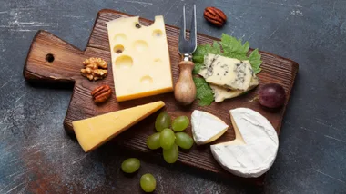 Cheese board with various cheeses, grapes, nuts, and a cheese fork, presented on a rustic wooden board.