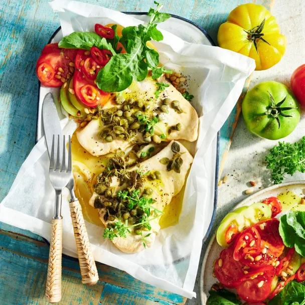 BBQ fish cooked in paper on blue background with cutlery and tomato salad