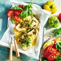 BBQ fish cooked in paper on blue background with cutlery and tomato salad