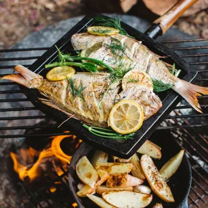 Whole fish and potato wedges cooking in pans over a campfire.