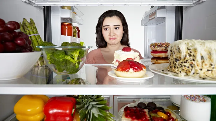 Women contemplates healthy versus unhealthy foods inside a fridge