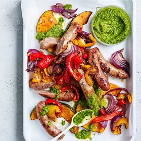 Cooked sausages and vegetables on a white rectangular platter with pesto dressing.