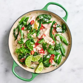 An aerial view of a large green pan filled with green beans, snow peas, and chicken in coconut sauce. Garnished with lime wedges, coriander and red chilli.